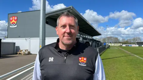 Tom Jackson/BBC Duncan Rumbelow smiling and looking at the camera. He is wearing a light jacket with 'DR' and the Cambridge City FC slogan on the chest. Behind him is the club's pitch at Sawston. 