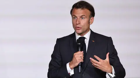 France's President Emmanuel Macron in a white shirt and suit holds a microphone and talks to an audience