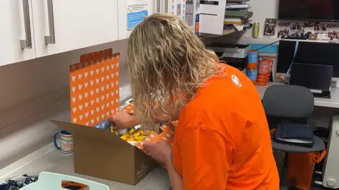 A woman in an orange T shirt packs a brown cardboard box with items. 