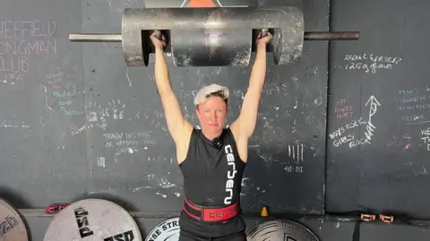 Laura Hollywood stands in a gym holding a large metal log bar overhead with both arms extended. She is wearing a black sleeveless top with white vertical text and a red lifting belt. Behind her is a dark wall with chalk writing and several large weight plates stacked on the floor.
