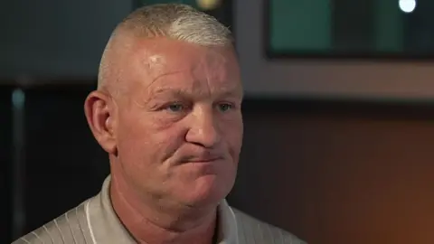 A head and shoulders photo of Dean Windass during a BBC interview. He has short, grey hair and is wearing a silver shirt.