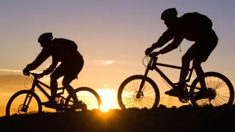 Two mountain bikers wearing helmets and ride bikes are silhouetted against an orange sunset. 