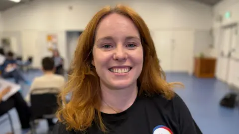 Steve Hubbard/BBC Kasia Truefitt has auburn shoulder-length hair and is smiling at the camera. She is wearing a black T-shirt with a Herts Inclusive Theatre badge.