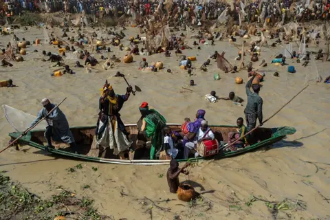 AFP via Getty Images A man dressed in traditional clothes rides on the front of a canoe surrounded by fishermen in the water.