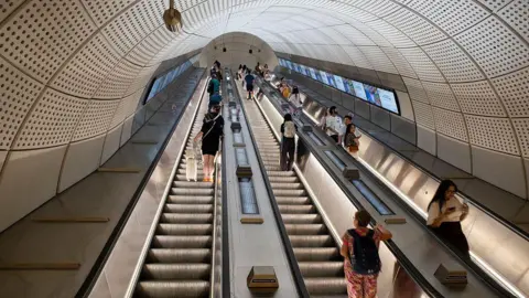 General view of a triple escalator at Liverpool Street station. Two are going up and one is going down. Various passengers are standing on all escalators.