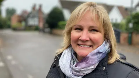 Amanda Mellor, smiling into the camera, has blonde hair and is wearing a light scarf and a dark jacket. The blurred background shows she is standing in a residential street.