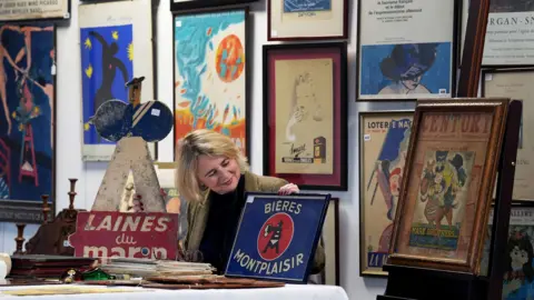 A woman sits at a table holding and looking at a enamel sign. All around her are prints and signs from the 20th century.