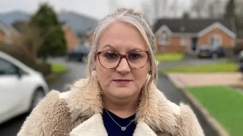 A woman wearing glasses and a fluffy beige and cream coat stands looking into the camera.