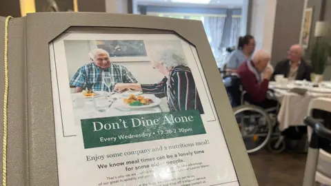 An invitation reading 'Don't Dine Alone' displayed in the restaurant area of a care home. In the background, a visitor, a care worker and a resident sit together at a table.