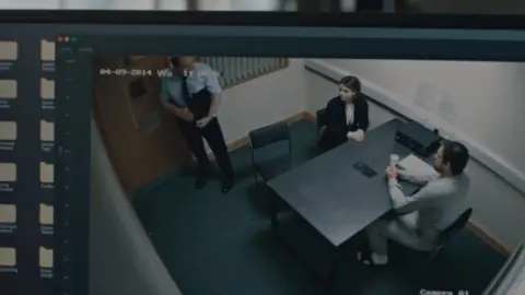 Camera footage viewed on a computer screen, showing a police interview room and three people around the table.