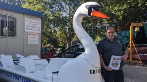 Alan Thornewill A man in his 60s smiles as he stands beside a swan-shaped pedalo boat, which is on land beside a portakabin at a boatyard. He has short hair and a short grey beard and is wearing a navy polo shirt, shorts and glasses. He is holding a poster.