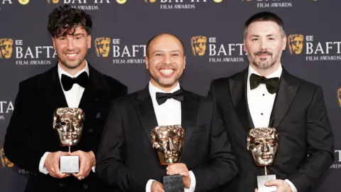 Three men stand on the 2026 BAFTA red carpet in black tuxedos. Each of the three hold a BAFTA award in the shape of a gold theatrical mask.