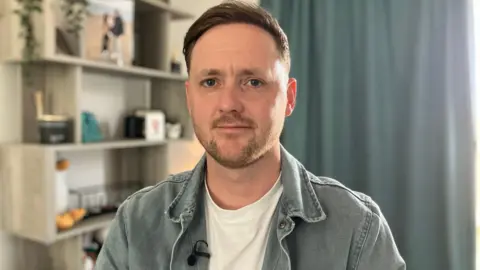 Chris Rodgers. He has short brown hair and facial hair. He is looking directly at the camera and smiling. He is sat inside a house with shelving, photo frames and a curtain in the background.