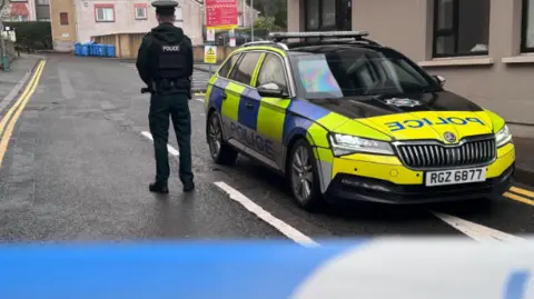 A police officer stands with his back to the camera beside a Skoda Superb estate police car. In the foreground directly in front of the camera is a blurred blue and white police tape.