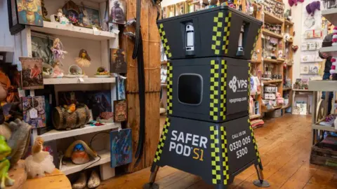 Northamptonshire OFPCC A tall black unit which is in three sections and widens at the top and bottom. It has yellow and black police stripes on the corners and "Safer Pod S1" written on the bottom segment. It is standing on the parquet floor of a shop which has many shelves displaying antique and collectable items.