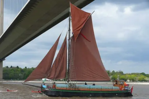 Patrick Hill Spider T sailing under the Humber Bridge, It has red-coloured sails and rigging. There is a person in a canoe nearby.