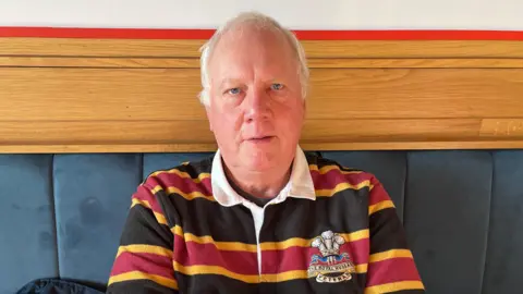 BBC/Julia Lewis A man with grey hair is wearing a blue, yellow and red striped rugby shirt.
