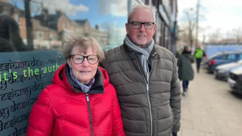 Jonny Humphries/BBC Dorothy Cummings, dengan mantel merah cerah, dan Bob Cummings, dalam mantel gelembung hijau, berdiri berdampingan di jalan yang sibuk tersenyum di kamera.