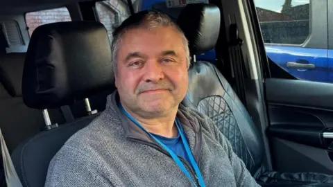 There is a man sat in a taxi with black leather interior seating. He has short grey hair, a grey stubbly beard, and a grey fleece jacket. He also is wearing a blue t-shirt and a blue lanyard string.