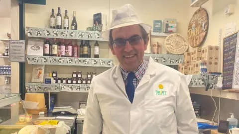 A man with a white hat and white coat, with a chequered shirt and blue tie underneath, stands in a shop. Behind him are three shelves of produce including jam and wine.