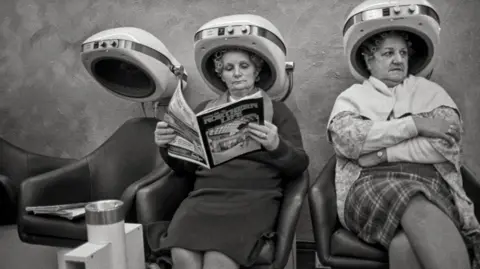 A black and white image of two women sitting in the hairdressers with heating cones on their heads. They are older women, one is reading a copy of Northern Life magazine and both have a rather cross expression on their faces 