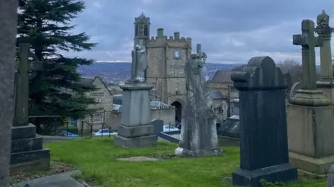 A graveyard in Sheffield. Several tombstones can be seen in the photo, surrounded by grass. The location has a high vantage point and looks across the rest of the city. 