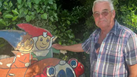 A man stands next to a plywood sign painted with the children's characters Noddy and Big Ears. He wears a short sleeve shirt.