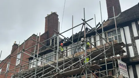 Jessica Richards Three men on scaffolding in front of a black and white timbered house with damage to its roof. They were green high-vis jackets and helmets. Behind them are several red brick chimney stacks.