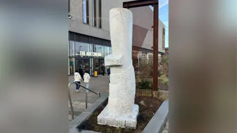 BBC A photograph of a white marble sculpture which looks like a tree stump. It is surrounded by bark on the floor and behind it plants can be seen. On the left, is the outside of a building with a sign that reads "The Arc Cinema".