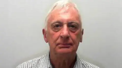 Man with white hair wearing a checked shirt looks into a camera.