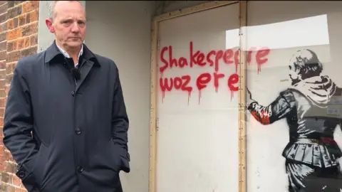 Clare Worden/BBC Lee Webster stands in front of the artwork in King's Lynn. It is painted on white double doors on a brick building. Art painted on white double doors on a brick building. It is an image in black of a character that looks like William Shakespeare from behind. He is holding a spray can, and the words "Shakespeare woz ere" have been written in red ink.