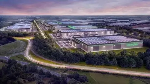 A large warehouse looking building lit up in the dark with Greenpower park written on the side in lit up green letters. A road runs in front and the buildings continue into the distance.