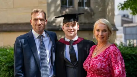 A family photo with two parents standing next to their son who is wearing a graduation robe. It is sunny and everyone is smiling.