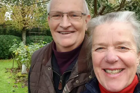 A selfie of Debs (taking the picture) and Simon. Debs has grey hair, tied back, and wears a blue coat and red polo neck. Simon has short white hair, and wears glasses and a brown coat.