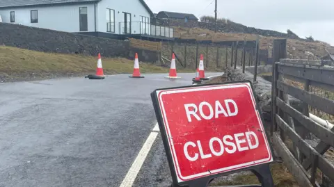 A red sign says "road closed" and red and white cones block access to the road. It is bordered by grassy verges which have yellowed, and one house with a glass balcony overlooks the road