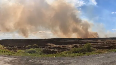A wide shot of a large plume of smoke over a mountainous area in Clogher. There are blue skies with some white clouds.