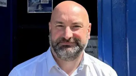 Marc Jones wearing a white shirt and smiling as he stands in front of a blue-coloured small booth. He is bald and has a dark-coloured, but greying beard. 
