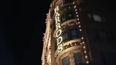 The exterior of Harrods department store, at night time, with the name of the store illuminated on the side of the building