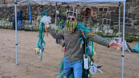 SAS A person standing on a sandy beach area under a white pop-up canopy. The canopy has printed text on the front that reads “SURFERS AGAINST SEWAGE” along with a web address beneath it. In the foreground, the central figure is dressed in a distinctive costume made from various pieces of plastic waste and debris. The costume includes strips of green material, plastic bags, and other items attached around the arms, torso, and legs. The person is also wearing sunglasses and a headpiece that incorporates similar materials, with strands hanging down over the face, partially covering it. One arm is extended outward, and the other hand holds a stick decorated with additional fabric, plastic, and a white flower-like object.