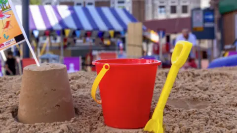 A sand castle next to a bucket and a yellow spade on a beach in a town
