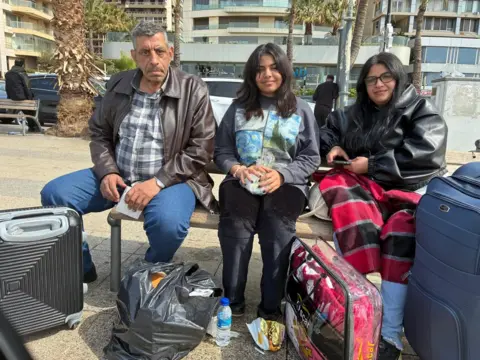 Dunia and parents sit on bench surrounded by luggage. 