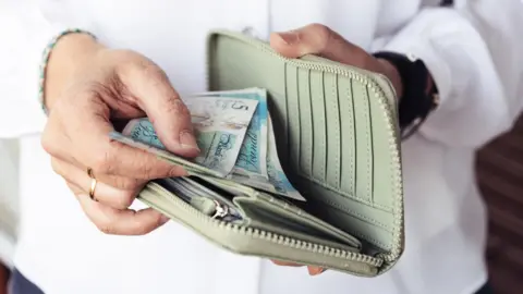 Getty Images Close up of hand of woman wearing a white shirt taking out five pound blue banknotes from her green purse
