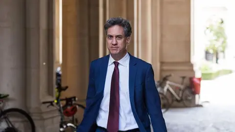 Getty Images Ed Miliband walking down a cobbled pedestrianised street in London. He is wearing a white shirt, blue suit, and red tie.