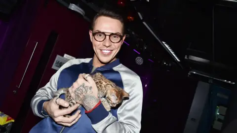 A man wearing black-rimmed glasses holds a kitten. He is wearing a shiny silver and blue jacket and is pictured against a dark background.