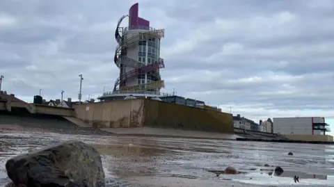 Redcar seafront with the vertical pier in the centre of the picture. The beach is wet and reflecting the sea wall.