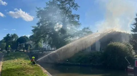 Claire Elizabeth Langridge A firefighter is using a hose to spray the smouldering pumping house from the other side of the canal