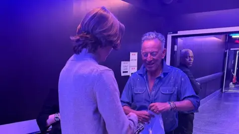 A teenage boy wearing a grey jumper stands facing Bruce Springsteen who is smiling. The boy is handing Springsteen a t-shirt. 