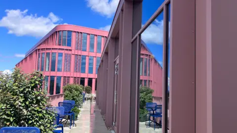 A view from the Balcony of the Forum. There are several blue chairs and bushes on the balcony, with some large windows on the right side. A red building with tall glass windows can be seen nearby.