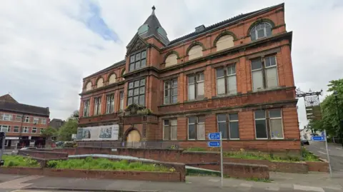 A street view image of a three-storey, 19th Century library building which has brick walls and greenery in front of it. Several windows are boarded up or broken.