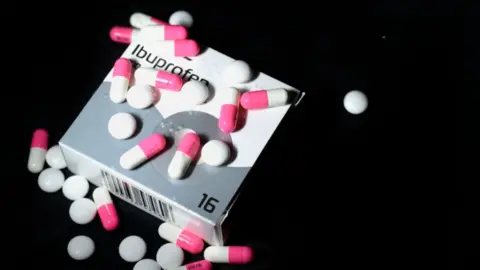 Box of ibuprofen with red and pink capsules and white tablets sprinkled on top.